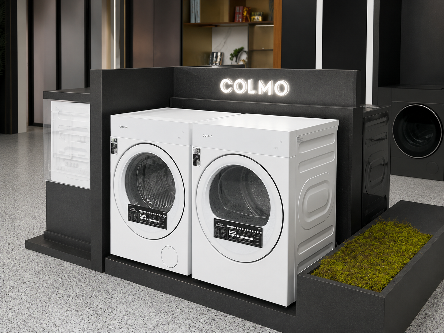 Angled showroom view of complete COLMO laundry appliance island display with two white appliances, illuminated sign, transparent information module and planted side trough
