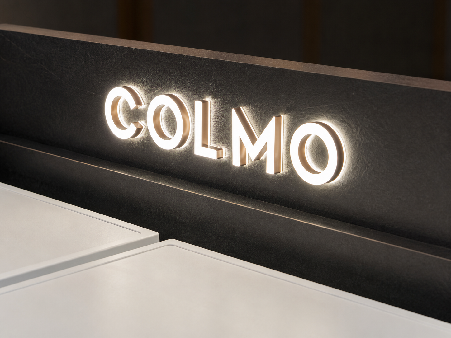 Close-up of illuminated COLMO lettering mounted on dark textured architectural panel